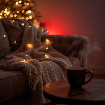 Cozy holiday living room at night with soft warm Christmas lights, a blanket on the couch, a mug of tea on a table, and subtle red ambient glow in the background. Peaceful, calm, relaxing, minimal aesthetic. No people. No therapy devices. Soft cinematic l
